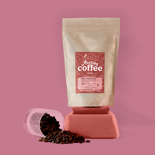 Pink background with a glass of coffee tipped over spilling beans. A bag of coffee rests on top of a pedestal with a red-orange label that has the Awesome Coffee Club logo. 