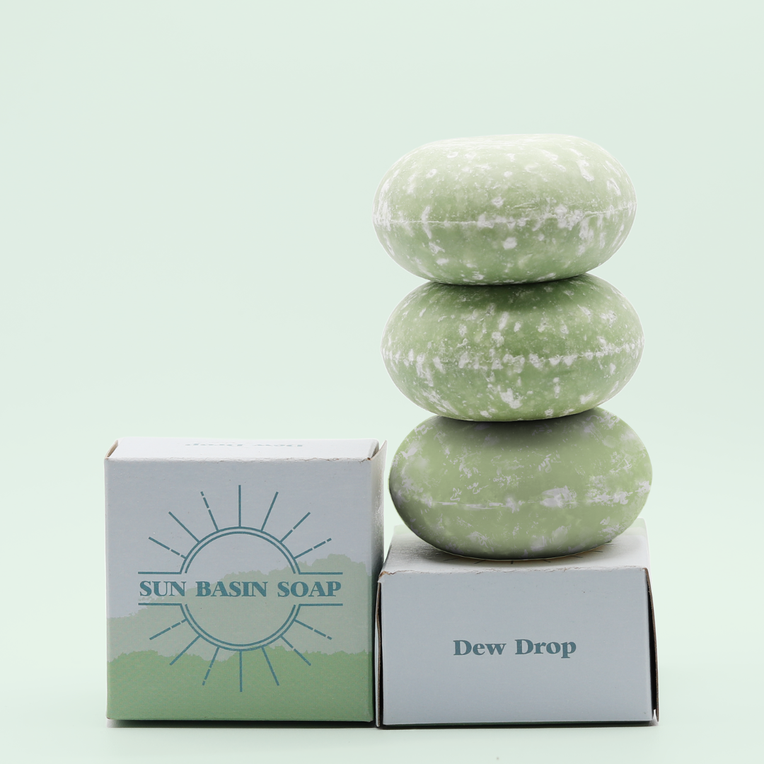 A stack of three of the Dew Drop Shampoo Bars on top of a small, blue box that reads Dew Drop. There is a small box next to it with the Sun Basin Soap logo in blue. A Good Store product.