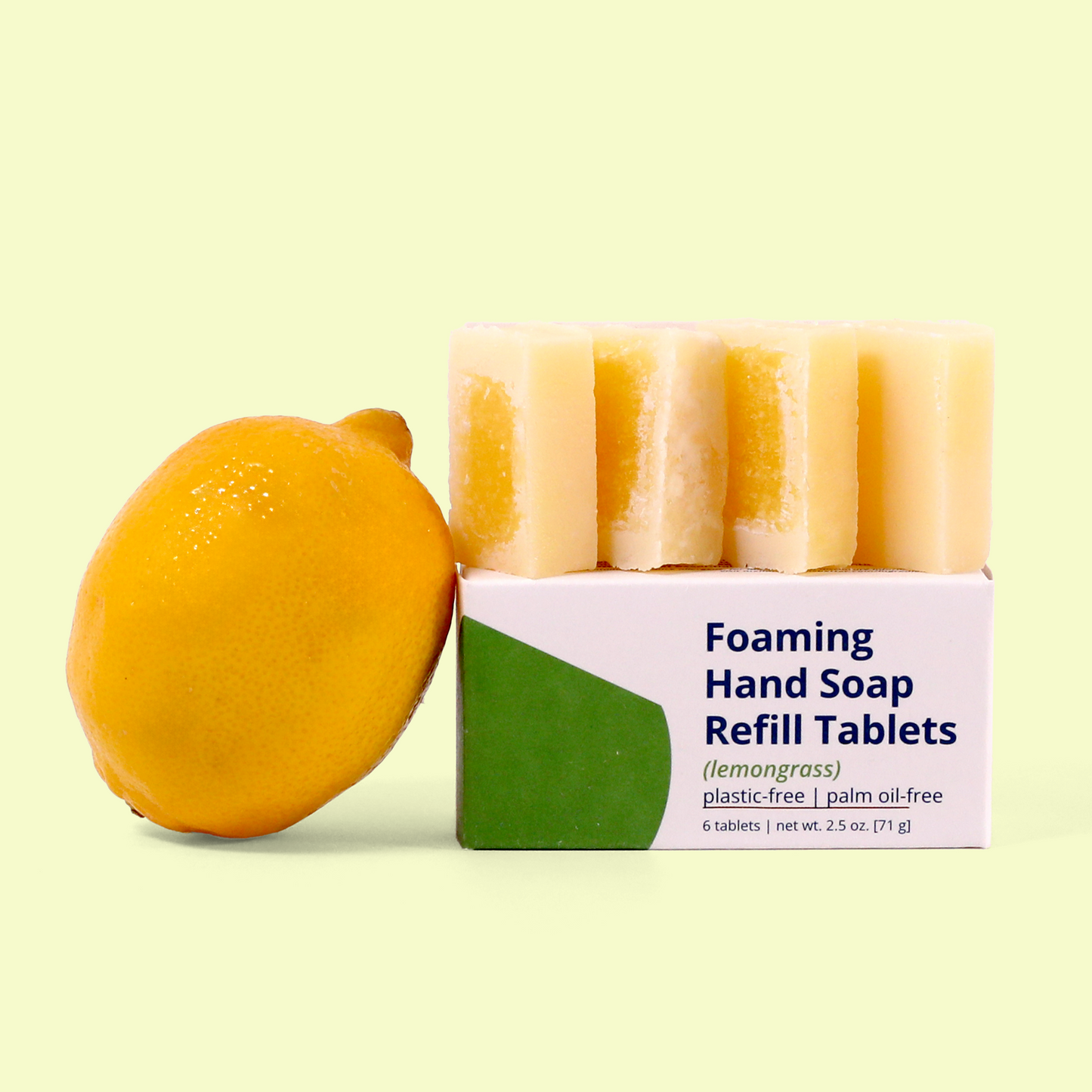 A box of Meliora lemongrass scented Foaming Hand Soap Refill Tablets with a lemon placed beside it against a pale yellow background. The white and green packaging specifies "plastic-free | palm oil-free" and "6 tablets | net wt. 2.5 oz. (71g)." Five light yellow soap tablets with darker yellow centers are displayed on top of the box. Foaming hand soap refill tablets from EcoGeek a Good Store product.