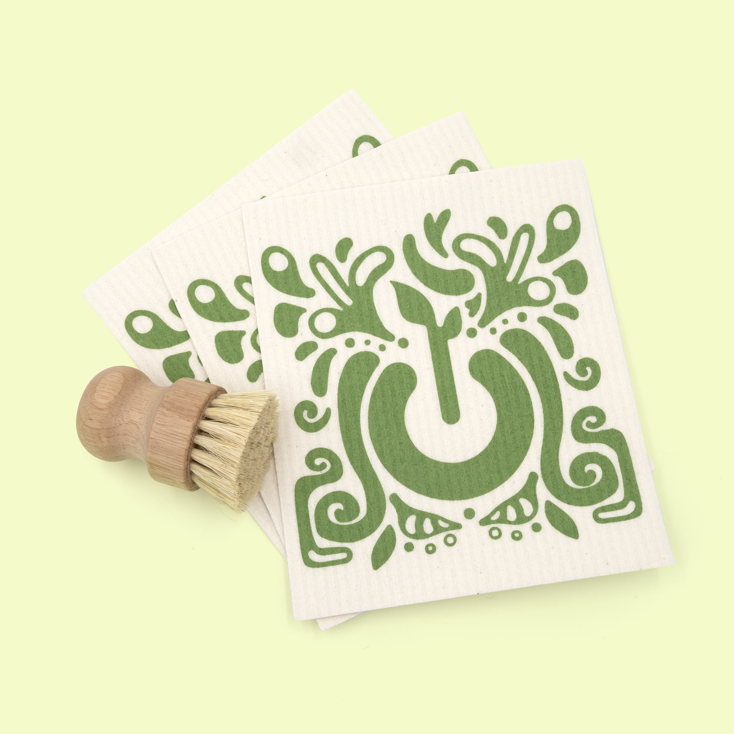 Three white square dish cloths with the same green tree design, displayed with a wooden cleaning brush with natural bristles positioned in the lower left corner. The items are arranged on a pale yellow background.