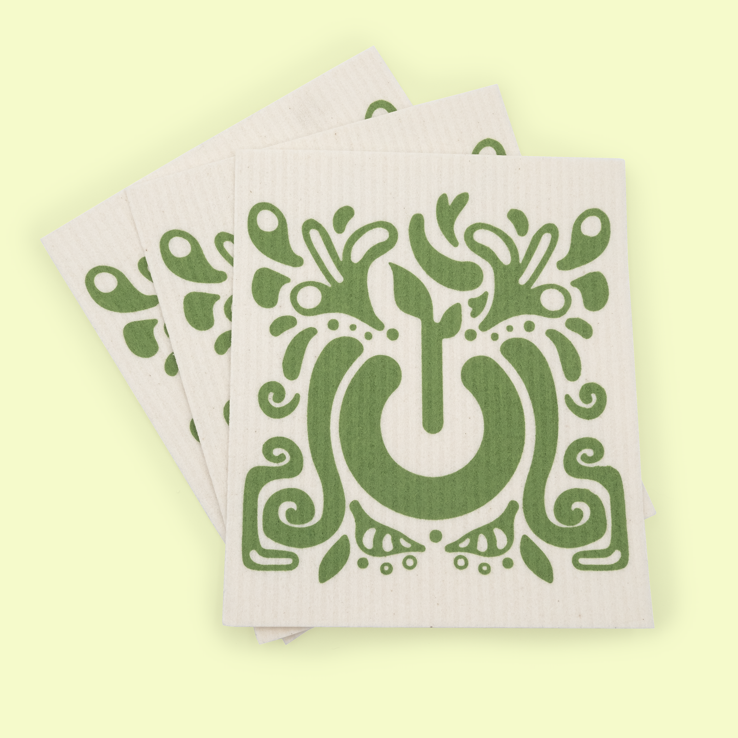 A stack of multiple dish cloths featuring the decorative green tree design, fanned out to show several copies. The botanical pattern displays an artistic tree with ornamental swirls, leaves, and flourishes in a folk art style, all set against a pale yellow background.