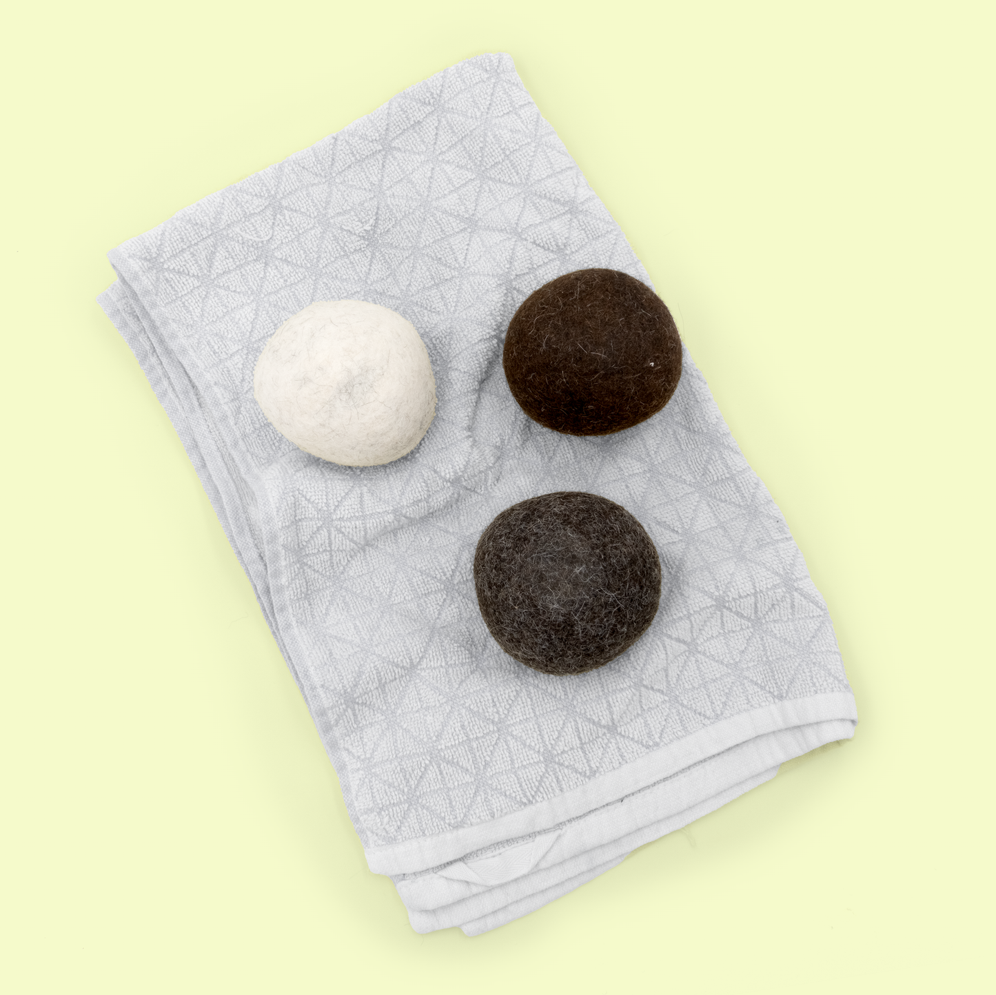 Three alpaca wool dryer balls arranged on a folded light gray linen towel against a pale yellow background. The balls consist of one white/cream colored ball and two dark brown balls, all showing the characteristic fuzzy, felted wool texture used for natural fabric softening in the dryer.