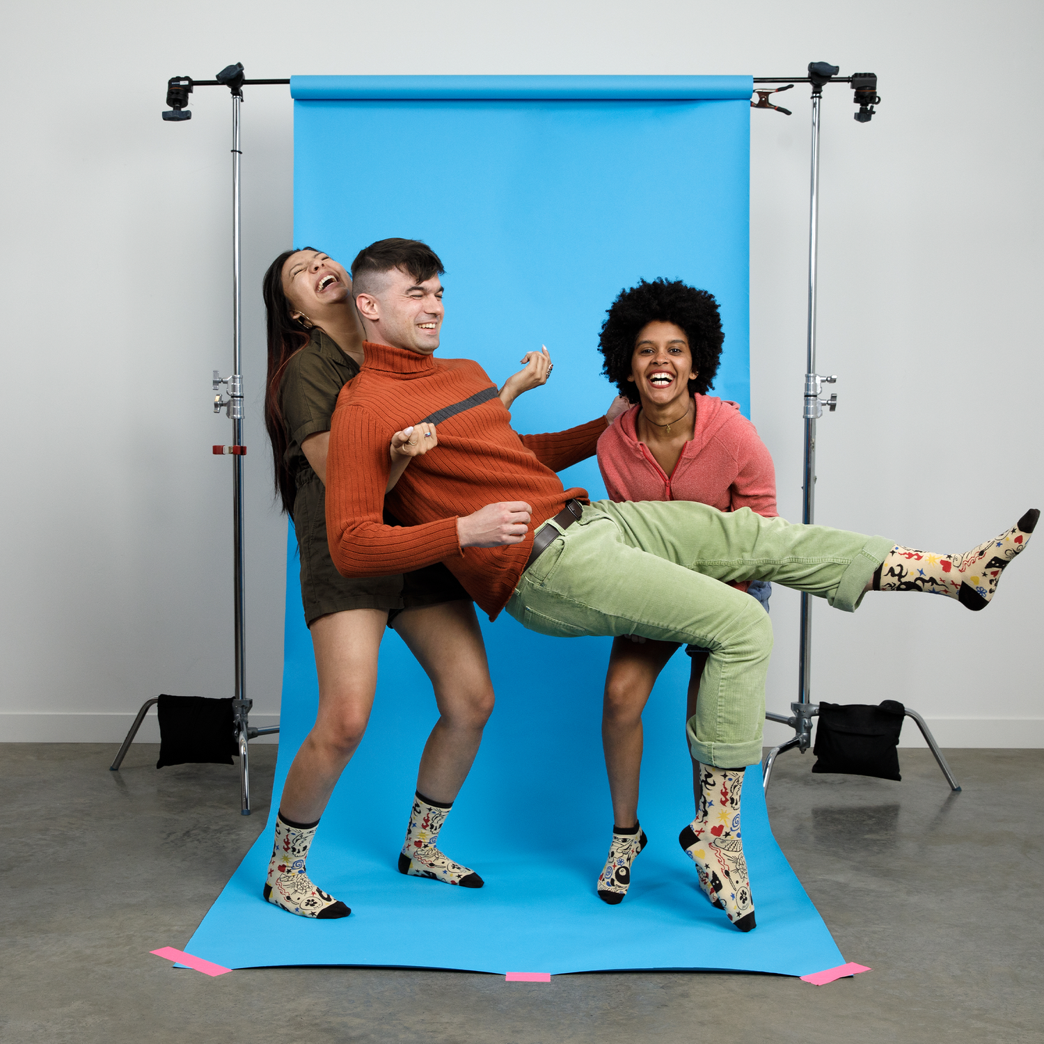 Three friends posing playfully against a bright blue photography backdrop. They're all wearing matching patterned Awesome Socks and laughing heartily. One person wears an orange turtleneck sweater and green pants, another wears a dark top, and the third wears a pink hoodie. They're arranged in a dynamic pose suggesting movement and joy.