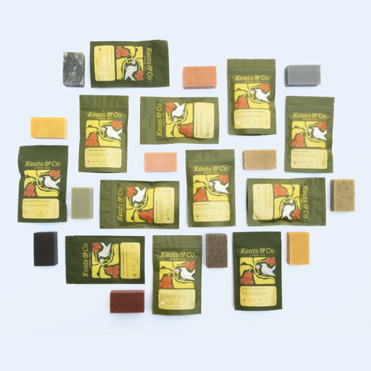A circular arrangement of Keats & Co tea packages and colorful soap bars on a light blue background. Green tea pouches featuring the brand's white bird logo alternate with rectangular soaps in various colors (yellow, orange, black, brown, gray). The composition creates a balanced display showcasing the complementary tea and soap collection, with visible varieties including "Glowing Gold Chamomile" and "Moonlit Mint."