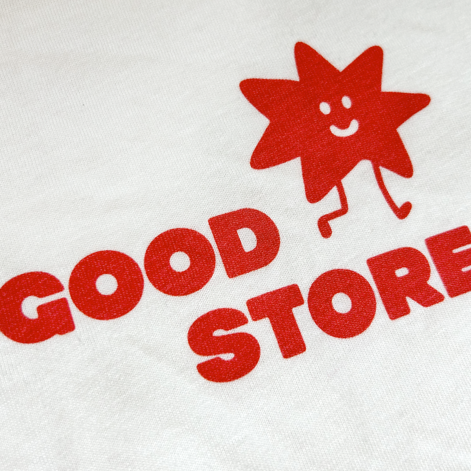 An extreme close-up of the red "GOOD STORE" logo on the beige fabric, clearly showing the smiling star character mascot with simple dot eyes and a curved smile, positioned above the bold red lettering.