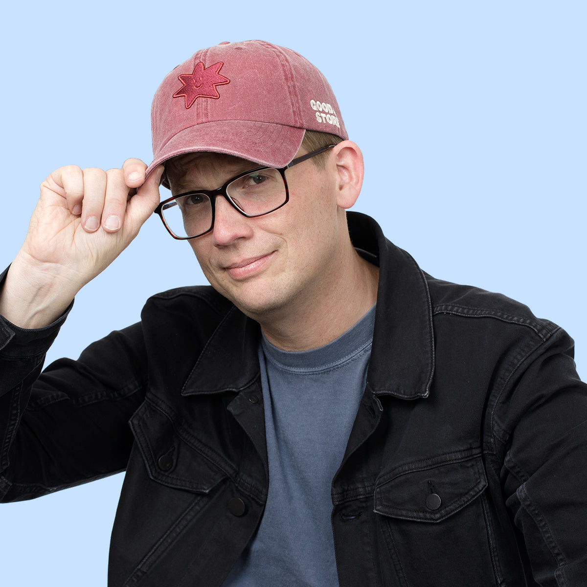 A man wearing a dusty rose red baseball cap with a red embroidered star and 'GOOD STORE' text, black glasses, a black denim jacket, and a blue t-shirt, posing against a solid light blue background.