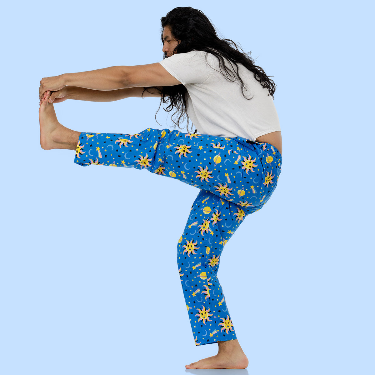 A human in a white shirt and blue pajama pants with a yellow sun and moon pattern poses in a yoga stretch against a light blue background.