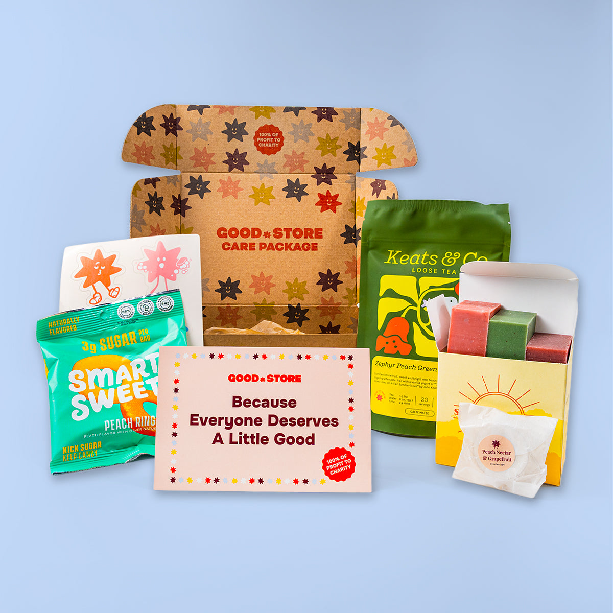 A curated gift set arranged on a light blue background, featuring a cardboard care package with star-patterned flaps, a green tea bag pouch, a bright green sugar packet, a pink welcome card, and a small white sachet.