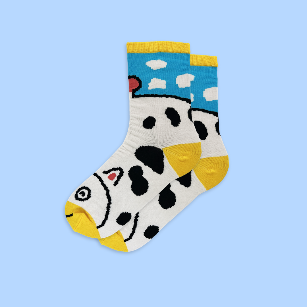 A pair of white crew socks with a playful cow pattern, featuring black spots, yellow toes and heels, and a blue sky section with white clouds at the top. The socks include cartoon cow faces with black and white eyes, pink inner ears, and black smile details.