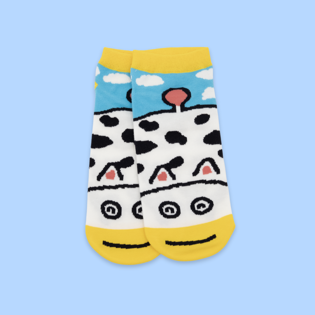 A pair of white ankle socks with the same cow-themed design - black spots throughout, yellow toe/heel sections, and a blue sky with white clouds at the top. Each sock features a cartoon cow face near the ankle with black outlined eyes, pink inner ears, and a black smile line.