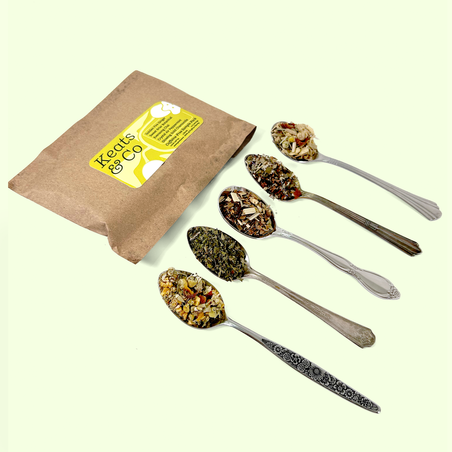 A brown kraft paper tea package with "Keats & Co" branding next to five silver spoons containing different loose tea blends. The teas appear to be herbal and fruit infusions with visible pieces of dried fruits, flowers, and tea leaves.