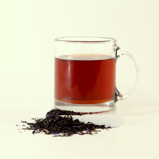 A clear glass mug filled with reddish-orange tea - Blue Fields Earl Grey -  next to a small pile of dried tea leaves. A Good Store product.