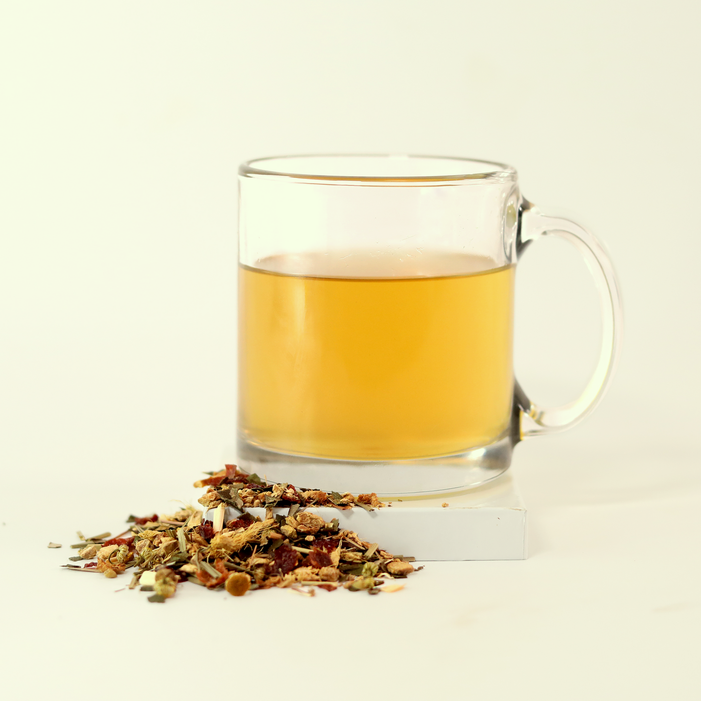 A clear glass mug filled with light yellow tea - Goldenfire Ginger -  next to a small pile of dried tea leaves. A Good Store product.