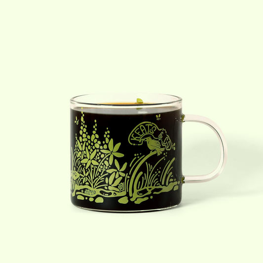 A detailed view of the Keats & Co Meadow glass mug. It's filled with coffee and the mug's design shows green illustrations of plants, flowers, and a bird, along with the Keats & Co logo. A Good Store product.