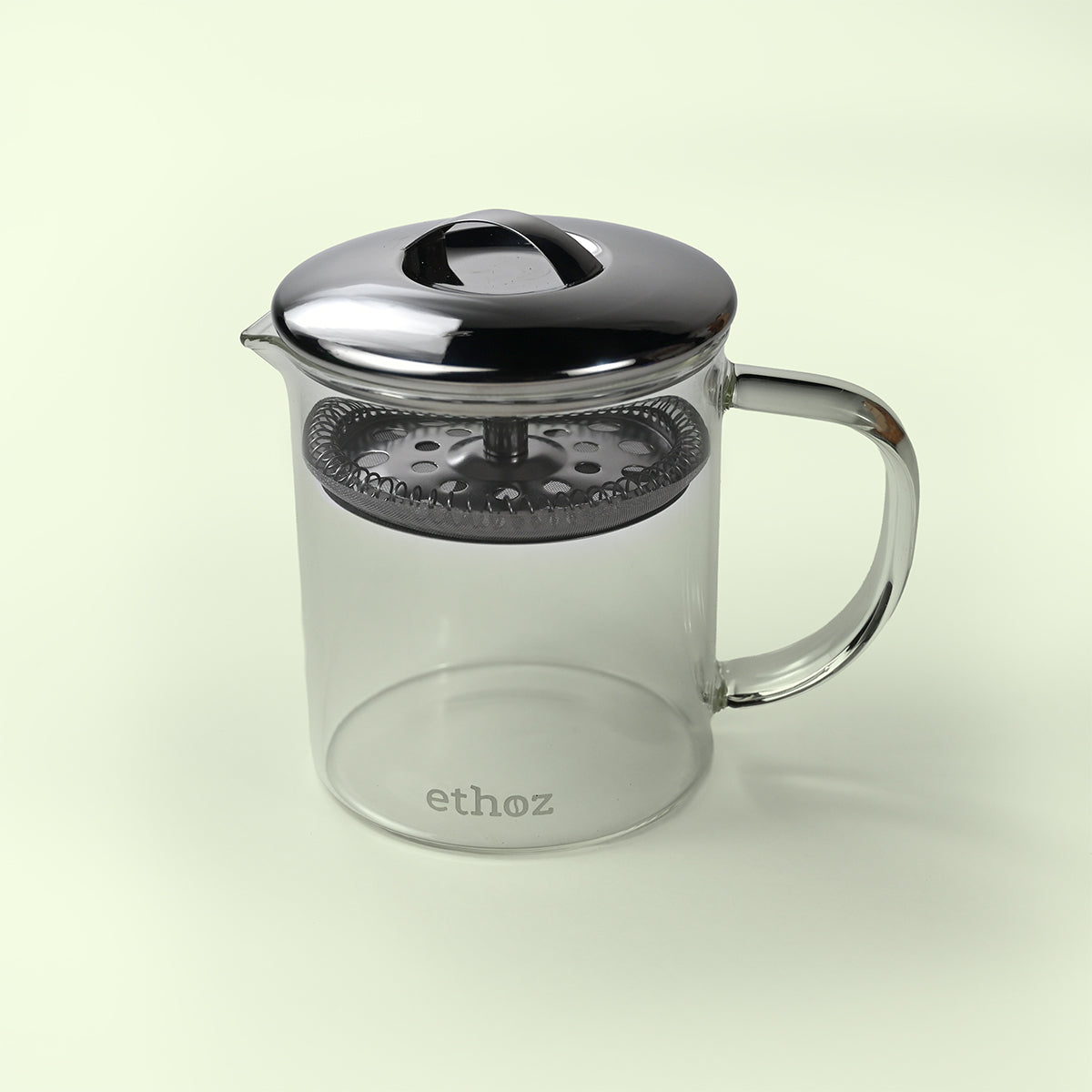 Clear glass tea infuser with metal lid and handle on a light beige background