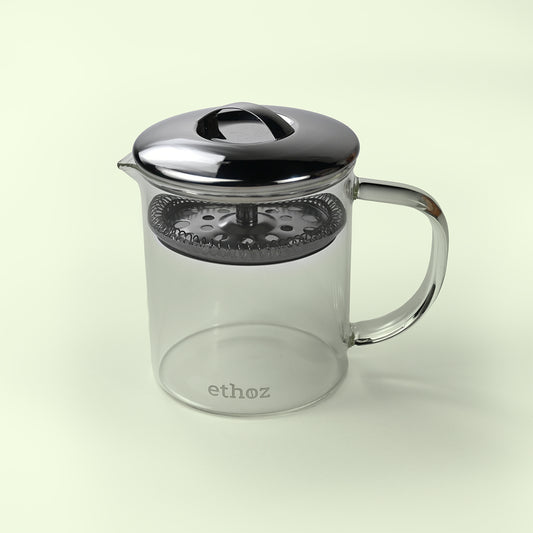 Clear glass tea infuser with metal lid and handle on a light beige background