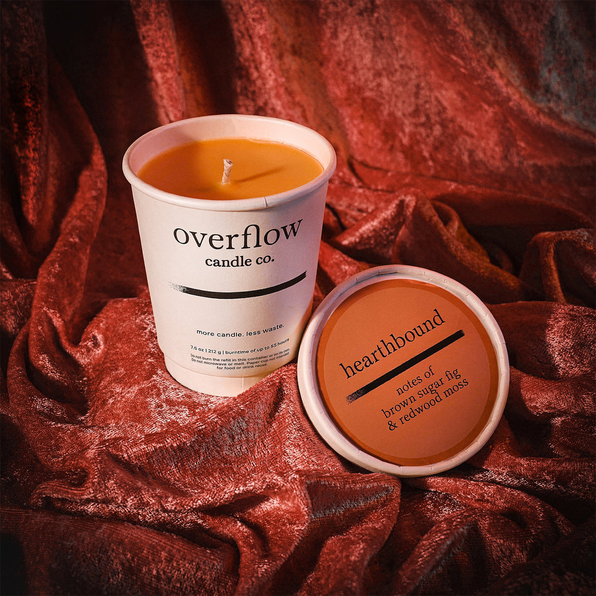 A white paper candle cup labeled 'overflow candle co.' with orange wax inside, shown beside its matching lid on draped rust-red fabric.