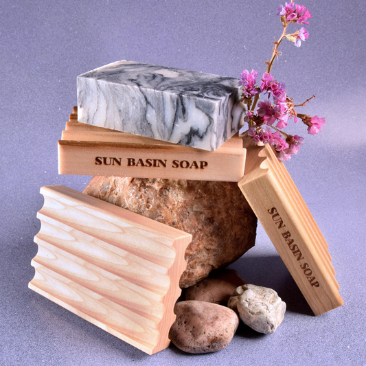 A white and black swirled bar of soap sits atop a ridged wooden soap saver dish that has Sun Basin Soap etched into the side. The soap saver rests on top of a rock with two others leaned against it. They are all in front of a lavender background. A Good Store subscription product.