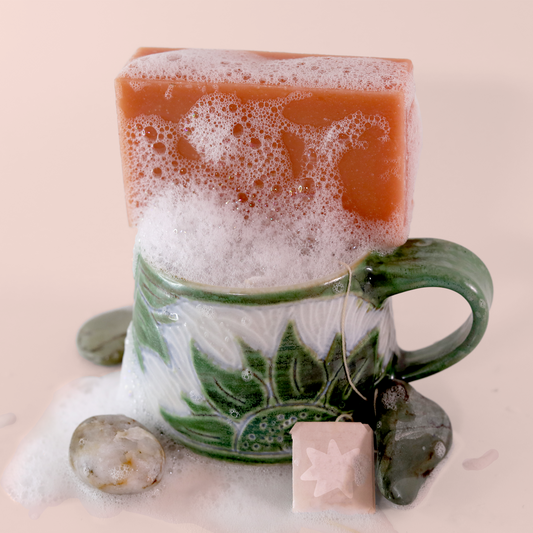 A bar of clay orange soap sits horizontally across the top of a small teacup. Foam and suds cling to the soap and fill the teacup to overflowing, dripping down its sides to pool on the ground. A tag for tea hangs out of the side of the teacup as well, on it is emblazoned a star shape, reminiscent of Dot, the Good Store's mascot. The background of the photo is very slightly orange.