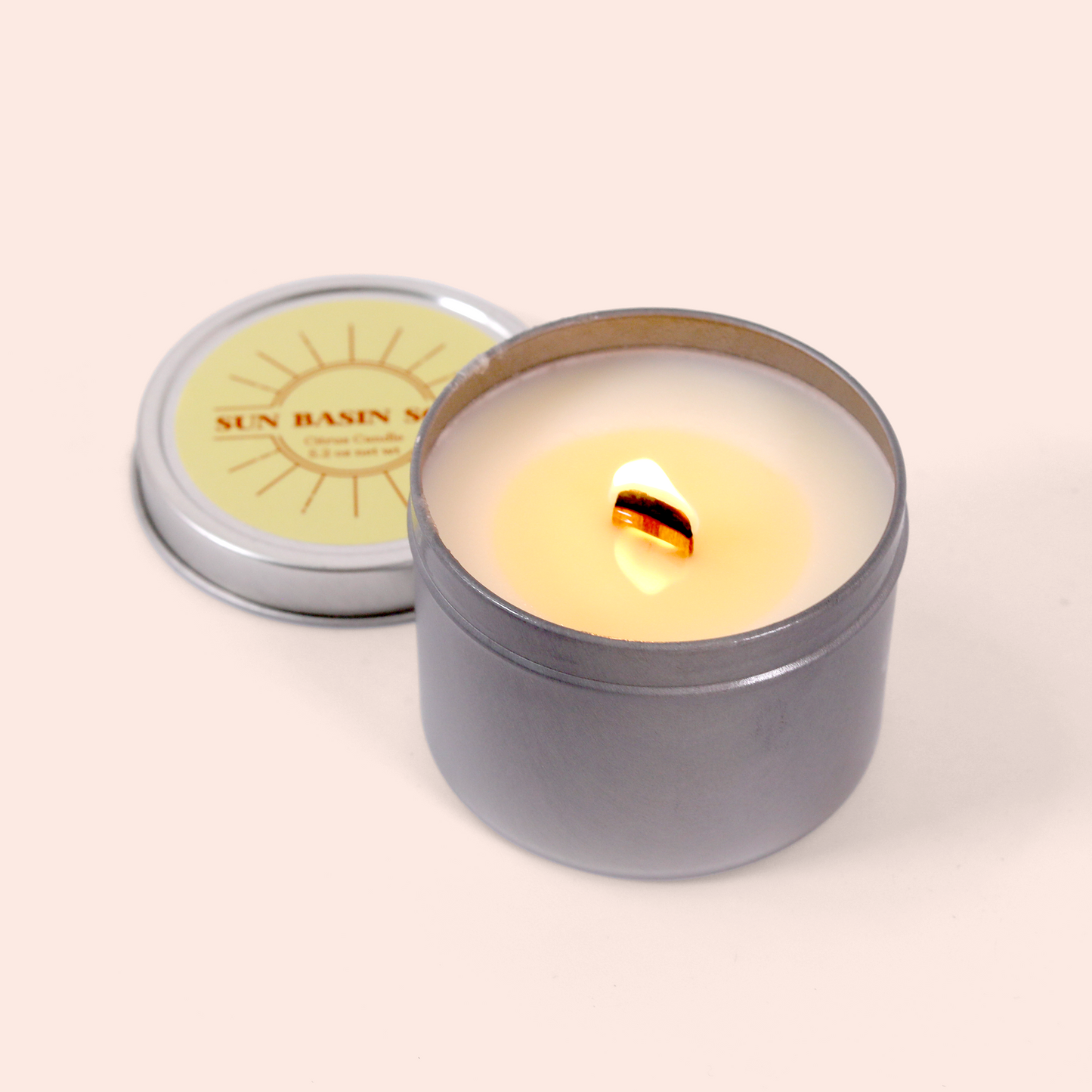 A lit candle in a gray metal tin container with cream-colored wax and a wooden wick,