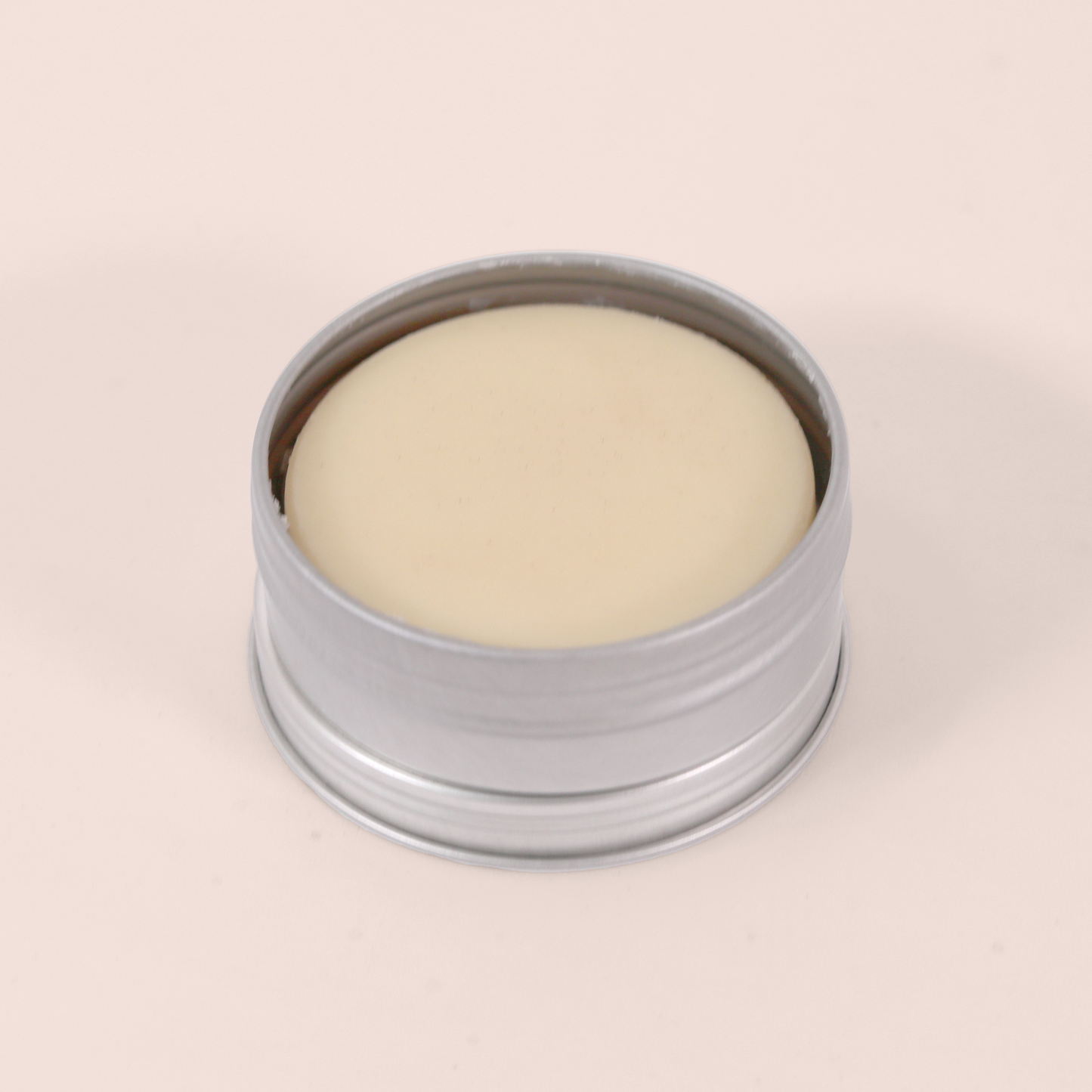 A single open silver tin containing a solid cream-colored lotion bar, photographed from an angled perspective against a light pink background.