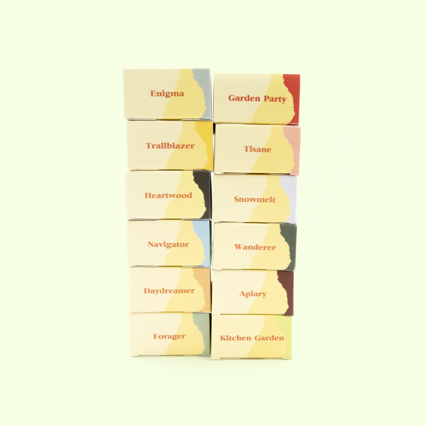A grid display of twelve small cream-colored boxes arranged in two vertical columns of six boxes each against a pale mint green background. Each box features a yellow abstract shape and is labeled with different names in orange text, including "Enigma," "Trailblazer," "Heartwood," "Navigator," "Daydreamer," "Forager" on the left column, and "Garden Party," "Tisane," "Snowmelt," "Wanderer," "Apiary," and "Kitchen Garden" on the right column. The boxes appear to be packaging for a collection of soap products