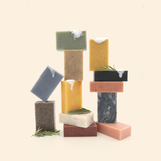 An artistic arrangement of handcrafted soap bars stacked in a balanced composition against a cream background. The soaps come in various colors including green, yellow, blue-gray, black, brown, white, pink, and marbled gray. Some bars have white lather or foam on their edges, and a few pieces are adorned with rosemary sprigs or sage leaves. The different colored bars are carefully balanced on top of each other to create a sculptural tower-like display.