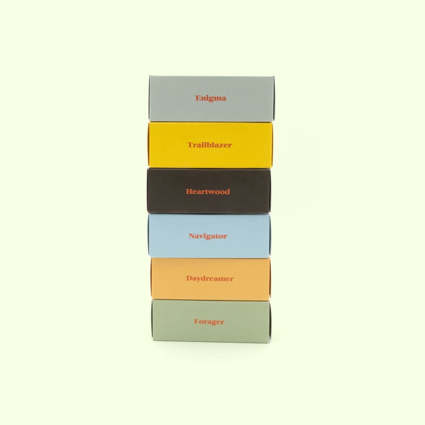 A stack of six colorful rectangular boxes arranged vertically against a light mint green background. From top to bottom, the boxes are labeled: "Enigma" (gray), "Trailblazer" (yellow), "Heartwood" (dark brown), "Navigator" (light blue), "Daydreamer" (peach), and "Forager" (sage green). Each box has its name printed in orange text.