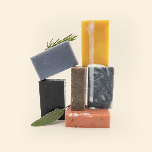 An artfully arranged collection of handmade soap bars in various colors and shapes against a cream background. The soaps include a blue-gray bar with rosemary sprigs, a black rectangular bar with a sage leaf, a brown speckled bar, a marbled gray and white bar, a yellow bar with some white foam, and a coral/pink rectangular bar at the bottom. The soap bars are stacked in a balanced composition.