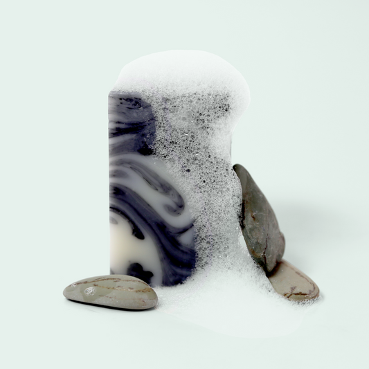 A rectangular block of marbled gray and white soap standing upright, partially covered in white soap lather. The foam appears to be cascading down one side of the soap. Three smooth, gray river stones are placed around the base of the soap block. The soap is set against a light blue background. Sun Basin Soap - Good Store.