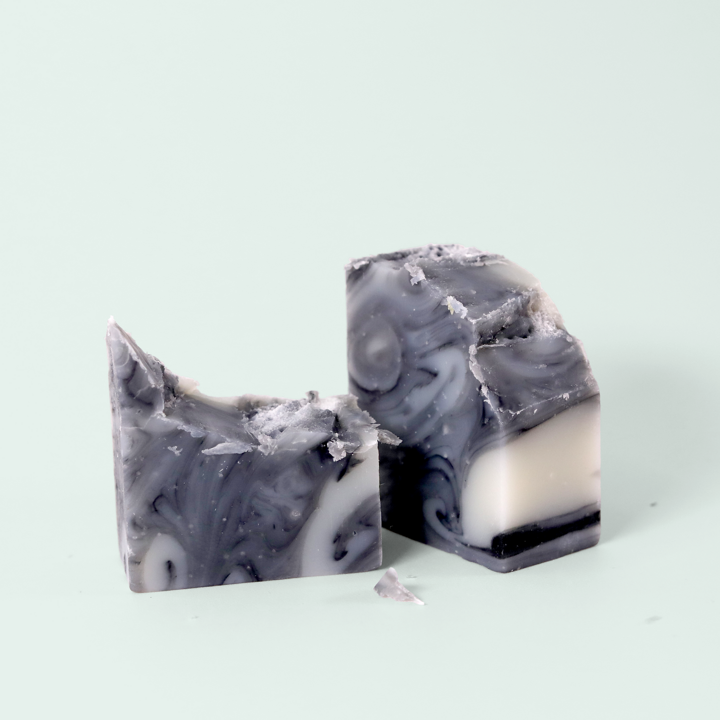 A marbled gray and white soap on a light blue background broken in half. Both blocks show a swirling pattern of dark and light gray with white streaks. The soap has a slightly rough, textured surface with some small flakes visible. Sun Basin Soap - Good Store.
