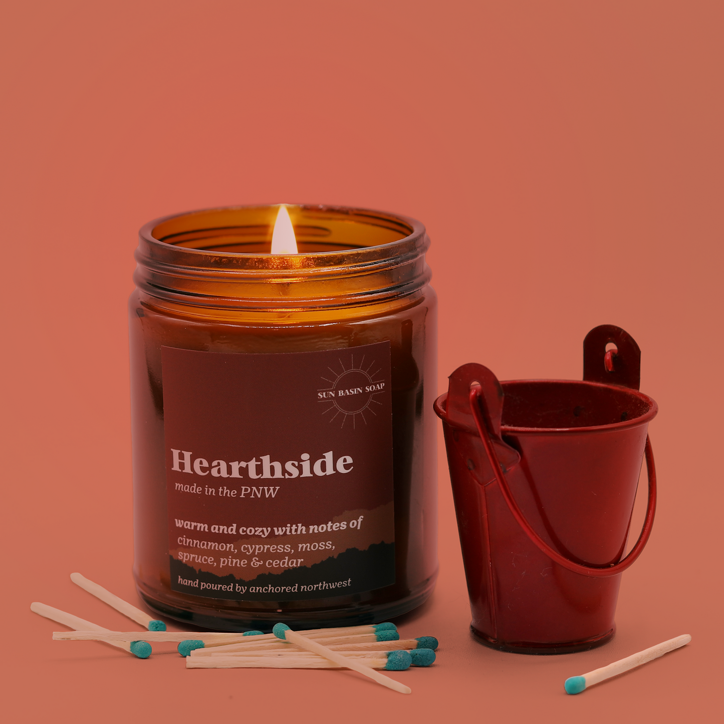 A candle in an amber jar with the text "Hearthside" and the Sun Basin Soap logo sits next to a little red tin, surrounded by matches. A Good Store product.