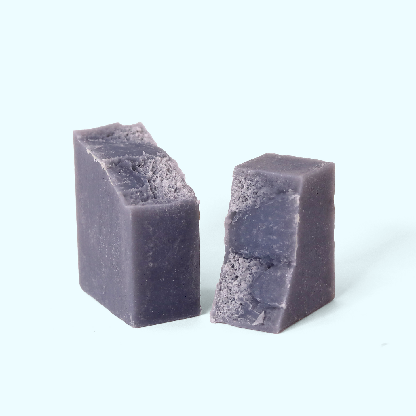 A rectangular block of purple-gray soap broken in half, on a light background. Both blocks have a rough, textured surface with visible grains or particles. The soap has a matte finish and a uniform color throughout. Sun Basin Soap - Good Store.