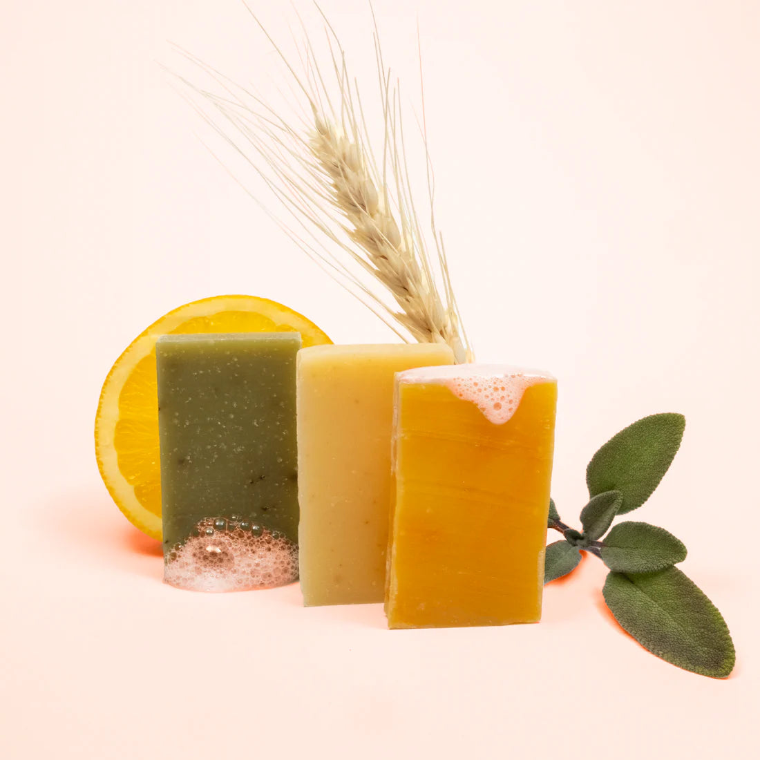 Three bars of soap in different colors with a lemon, wheat stalk, and leaf on a light pink background