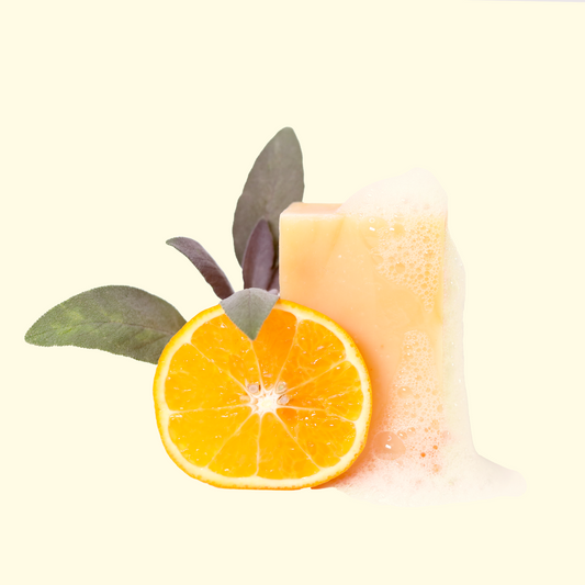 A pale yellow Trailblazer soap bar with foam, next to a sliced orange and herb leaves set on a light background. Sun Basin Soap - Good Store.