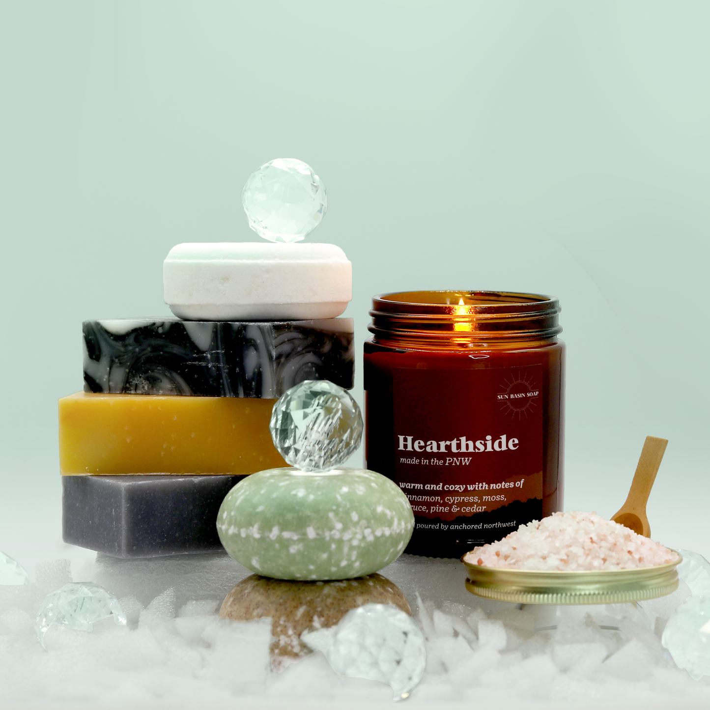 The Viridian Sun Bath Box from Sun Basin Soap contains a stack of three soap bars with a bath bomb resting atop. Two shampoo bars sit in front of a candle and bath salts. A Good Store product.