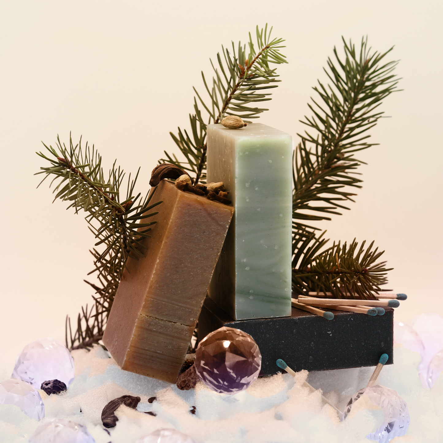Three holiday soaps from Sun Basin Soap, one brown, one light green and one black, are stacked together with sprigs of pine, cardamon and matches scattered around them. A Good Store product.