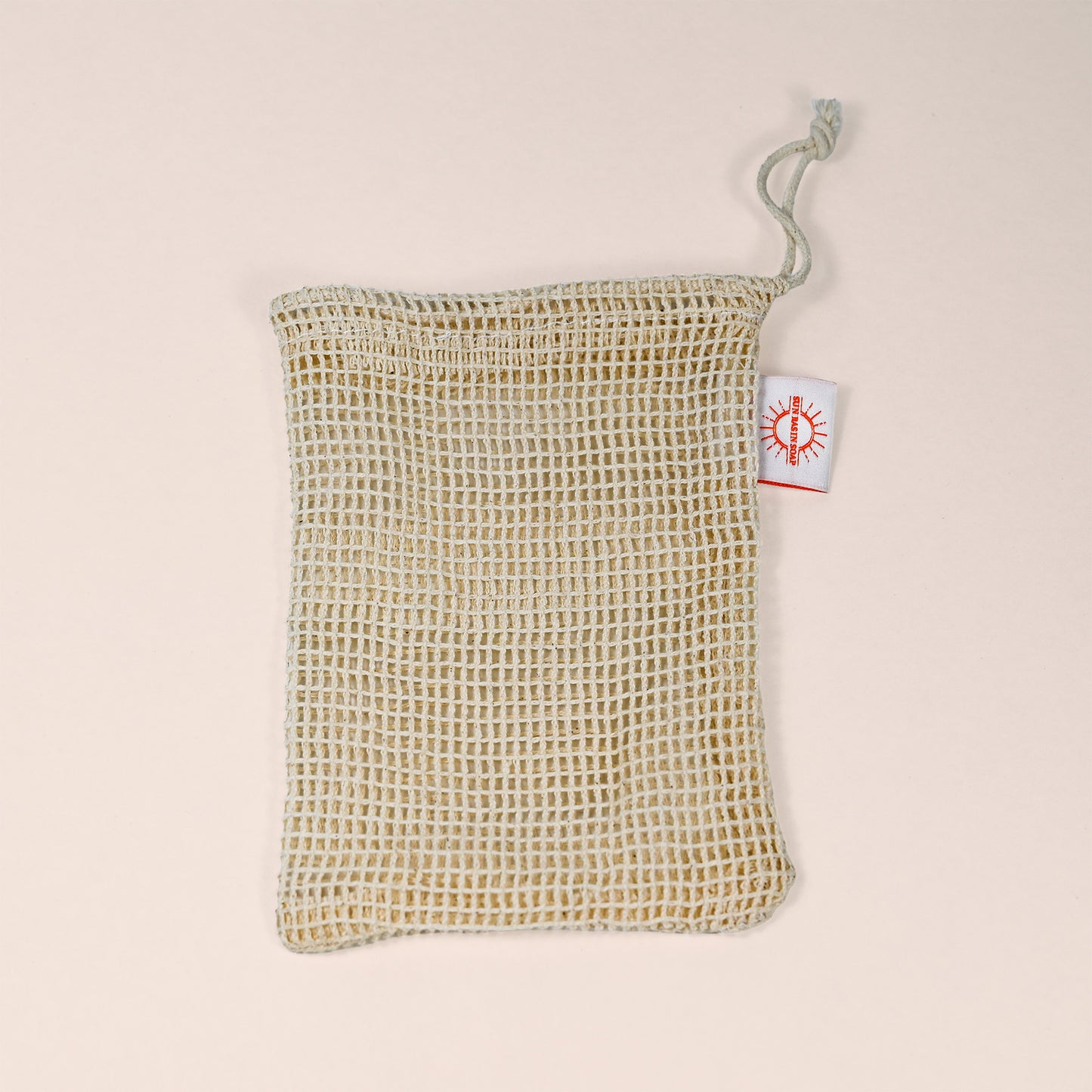 A natural sisal soap saver pouch with a drawstring closure and a small white tag featuring an orange sun-like logo, displayed on a light neutral background.