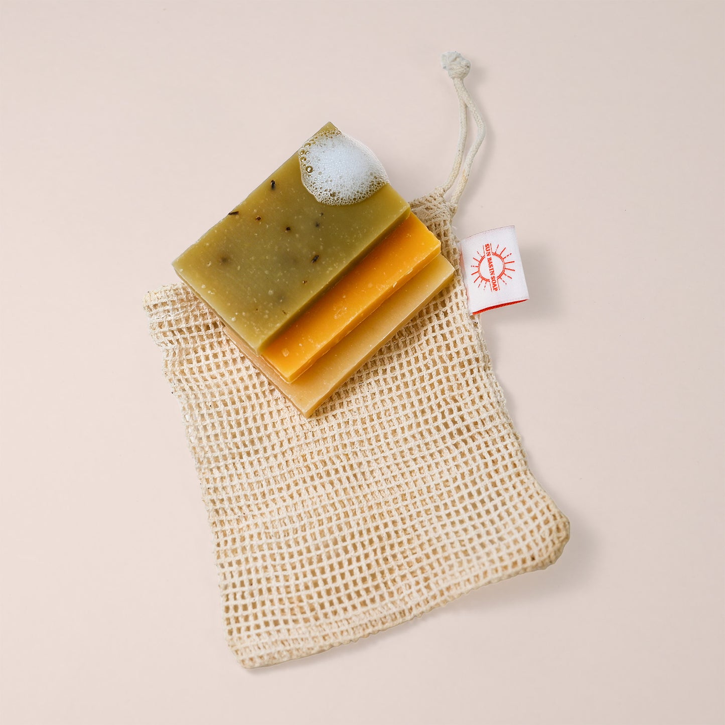 Three rectangular bar soap samples—green with speckles, golden yellow, and beige—rest on top of a woven sisal soap saver pouch with a white drawstring and orange sun logo tag, against a light beige background.