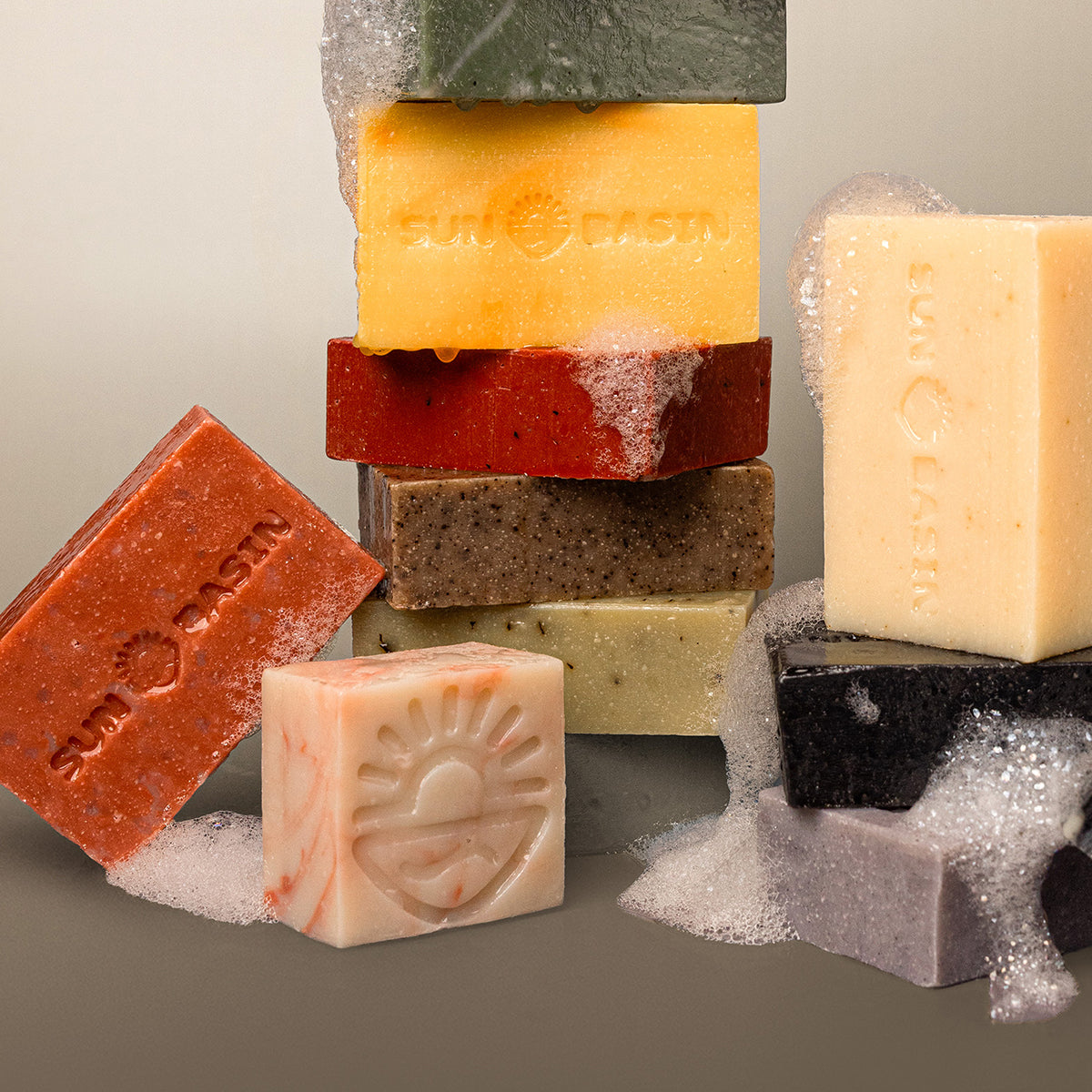 A styled product shot of multiple Sunbath soap bars in various colors, some stacked and standing upright, with white lather and foam around them on a neutral gray background.