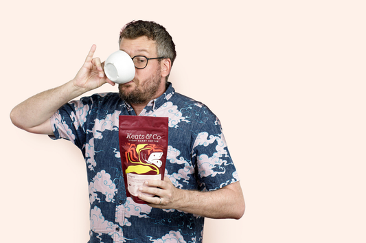 Image of John Green holding a bag of Keats & Co Coffee while sipping from a ceramic mug with his pinky up.