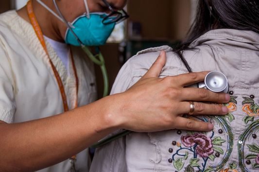Image of a person wearing scrubs and a medical mask listening to a patients breath with a stethoscope  