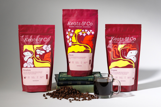 3 bags of Keats & Co coffee, one on top of a stack of books, next to a cup of black coffee and a pile of coffee beans