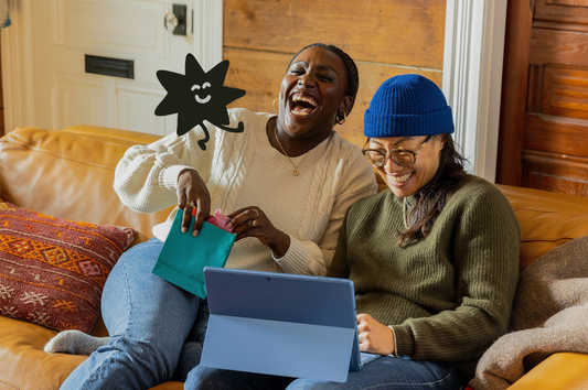 Two people laughing on a couch, one is looking at a tablet, the other is opening a gift. Dot, Good Store's mascot is on the image laughing along