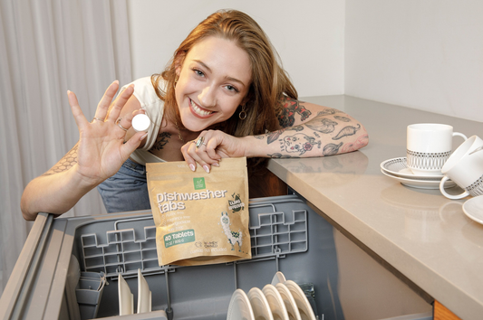A girl smiling at the camera in a kitchen holding a bag of Green Llama dishwasher tabs in one hand and a singular dishwasher tab in the other hand. She is leaning over a dishwasher filled with dishes