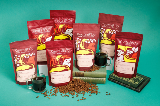 Image of a display of 7 bags of Keats & Co coffee on a table with spilled coffee beans, stacks of green books, and two glass cups of coffee.