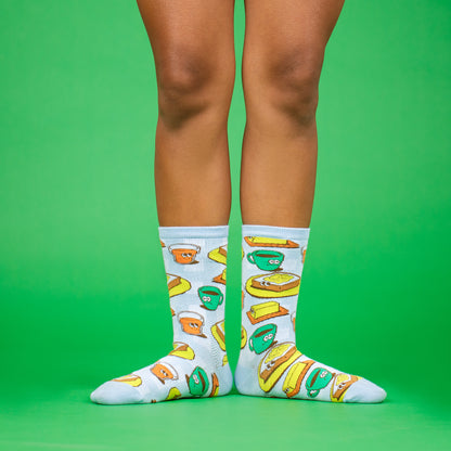 A green background showing two legs from the knee down in a ballet first position pose, wearing ankle socks from the Awesome Socks Club with colorful illustrations of toast, butter and coffee. A Good Store subscription product.