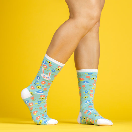 A pair of legs wearing Awesome Sock Club crew socks that are light teal with pastel flowers and bunnies on them. A Good Store product.
