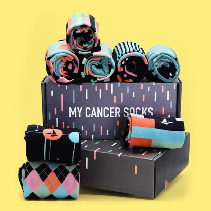 A stack of Hank's Cancer Socks in front of a yellow background atop black boxes with color dashes. The side of the box reads "My Cancer Socks". The socks are lined up and are differing patterns with similar color schemes. An Awesome Socks Club product.