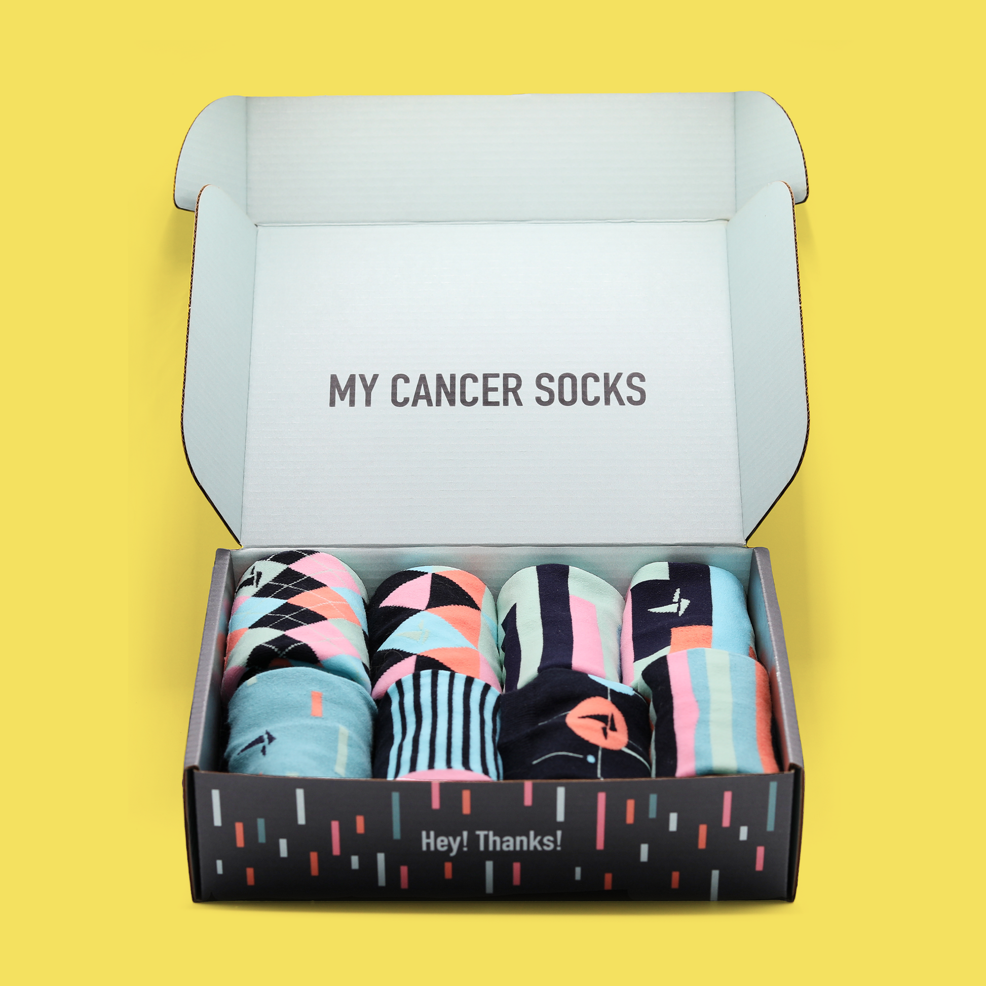 An open box of Hank's Cancer Socks in front of a yellow background. The inside of the box reads "My Cancer Socks". The socks lay in rows of two with four in each row and are differing patterns with similar color schemes. The side of the box reads "Hey! Thanks!". An Awesome Socks Club product.
